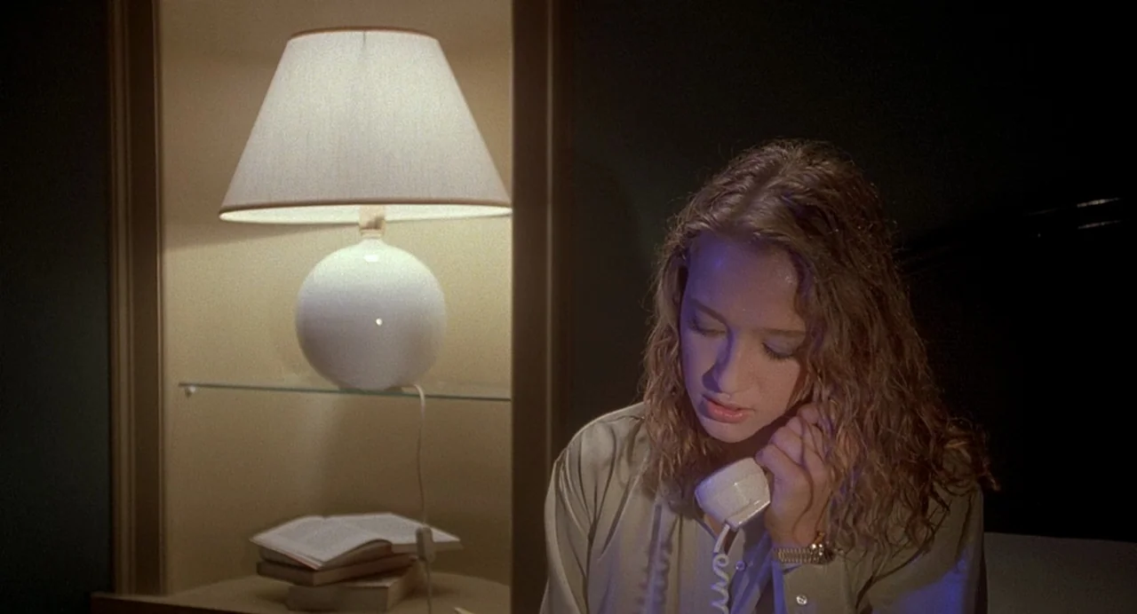 Inferno (1980) movie still: medium — A dark hotel room features a built-in shelf containing a lamp and books. A young woman sits in the foreground…