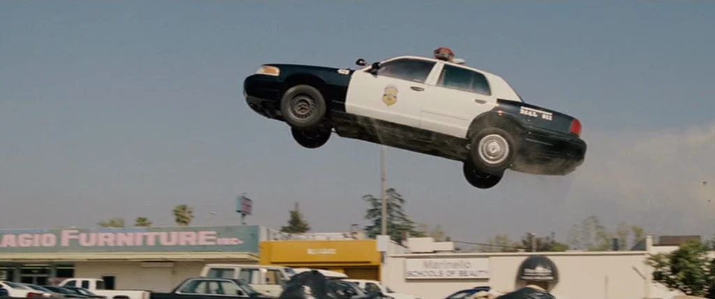 Pineapple Express (2008) movie still: wide — A suburban parking lot sits beneath a clear blue sky in front of businesses. A black and white police…