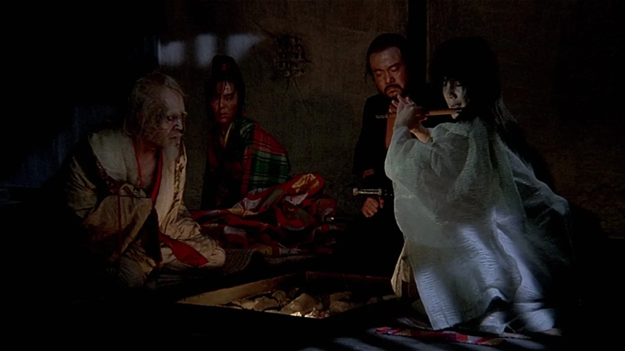 Ran (1985) movie still: medium — A dark, sparsely furnished room centers around a stone fire pit on the floor. Four figures sit on straw mats…