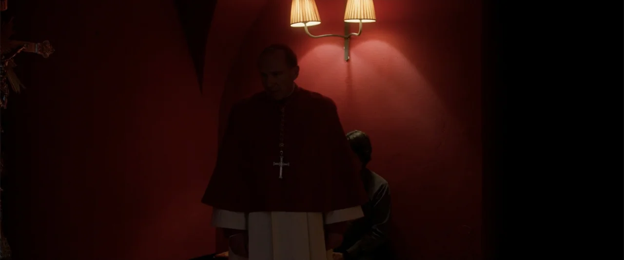 Conclave (2024) movie still: medium — A room with deep red walls features a wall-mounted double lamp emitting warm light. A man in religious…