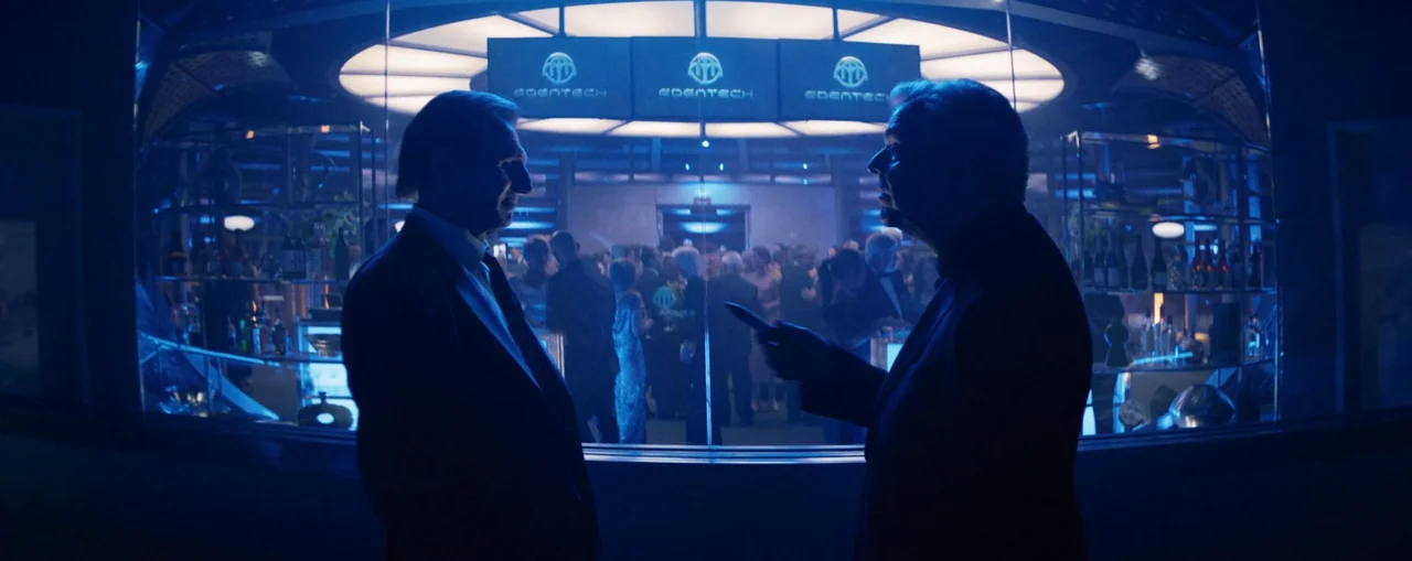 The Naked Gun (2025) movie still: medium — A high-end modern event space features glass walls separating an outer corridor from a crowd. Two men in…