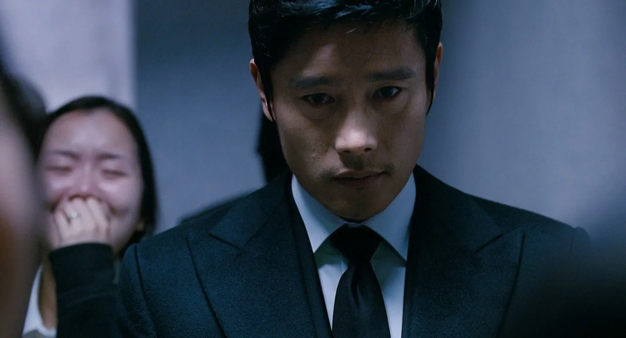 I Saw The Devil (2010) movie still: closeup — A dimly lit indoor space with cool gray walls and blurred surroundings. A man in a dark suit stares…