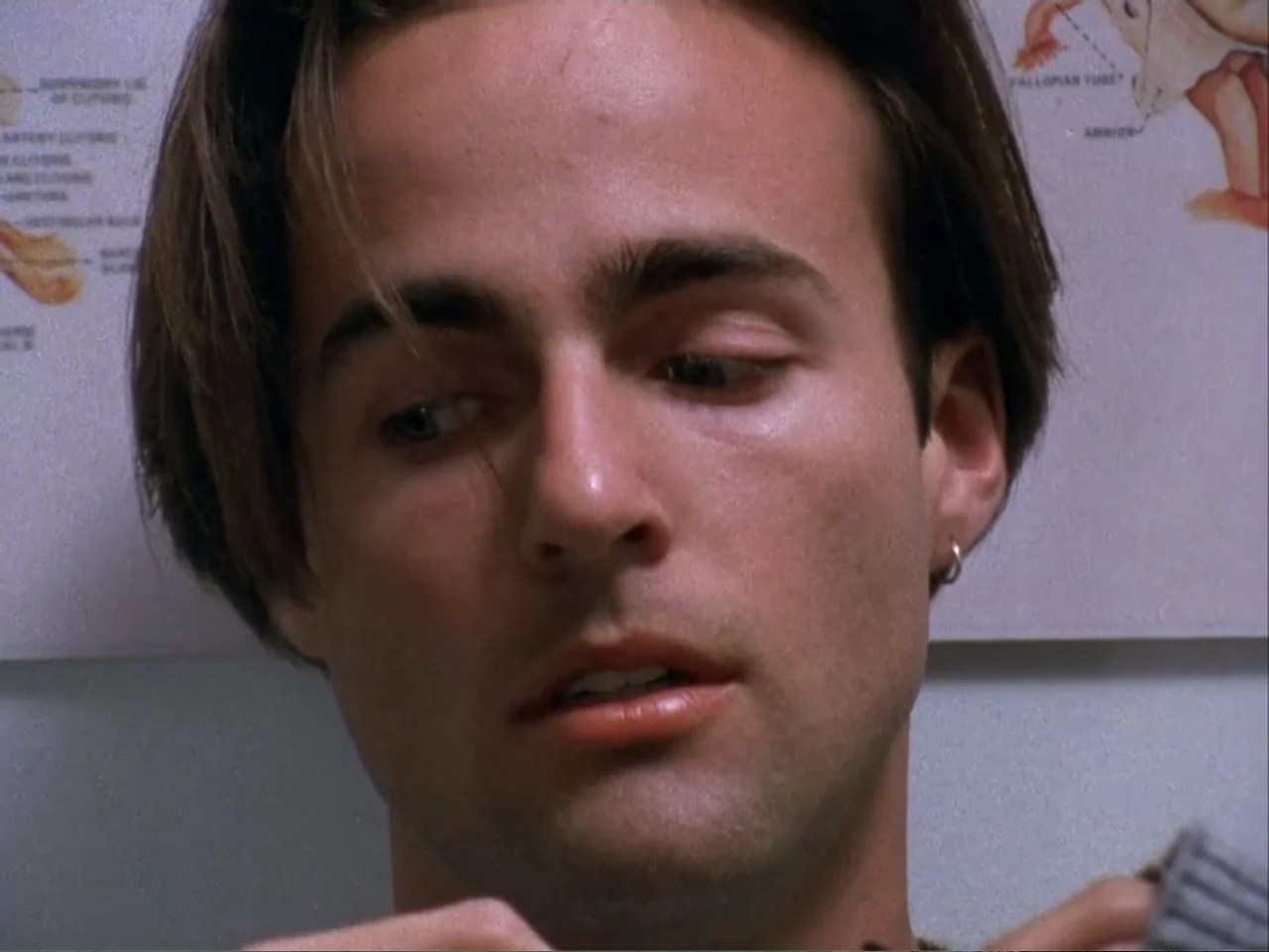The Living End (1992) movie still: closeup — A white wall with a medical diagram of the female reproductive system is visible behind. A young man…