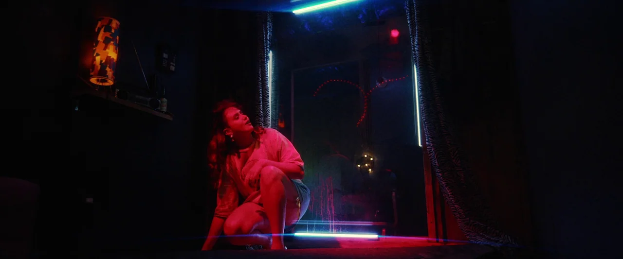 Revealer (2022) movie still: medium — A dark space lit by sharp cyan and magenta neon light tubes. A young woman crouches on the floor looking up…