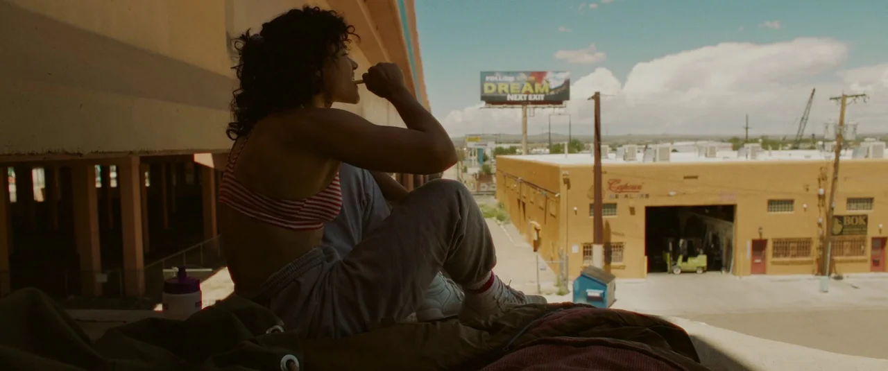Love Lies Bleeding (2024) movie still: medium — A woman sits on the concrete edge of an industrial overpass overlooking a street. A billboard…