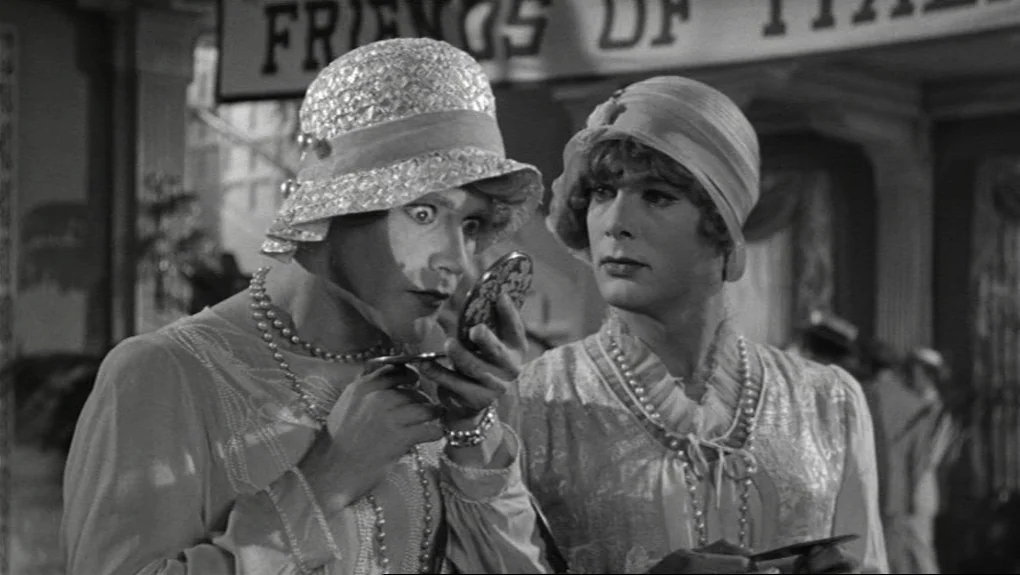 Some Like It Hot (1959) movie still: medium — Two men disguised in 1920s women's clothing stand indoors near a sign. One man inspects his reflection…