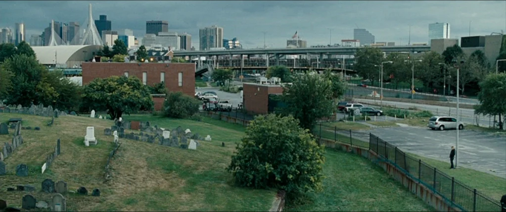 The Town (2010) movie still: establishing — A sprawling graveyard overlooks a urban parking lot and highway under a cloudy sky. The cityscape looms…