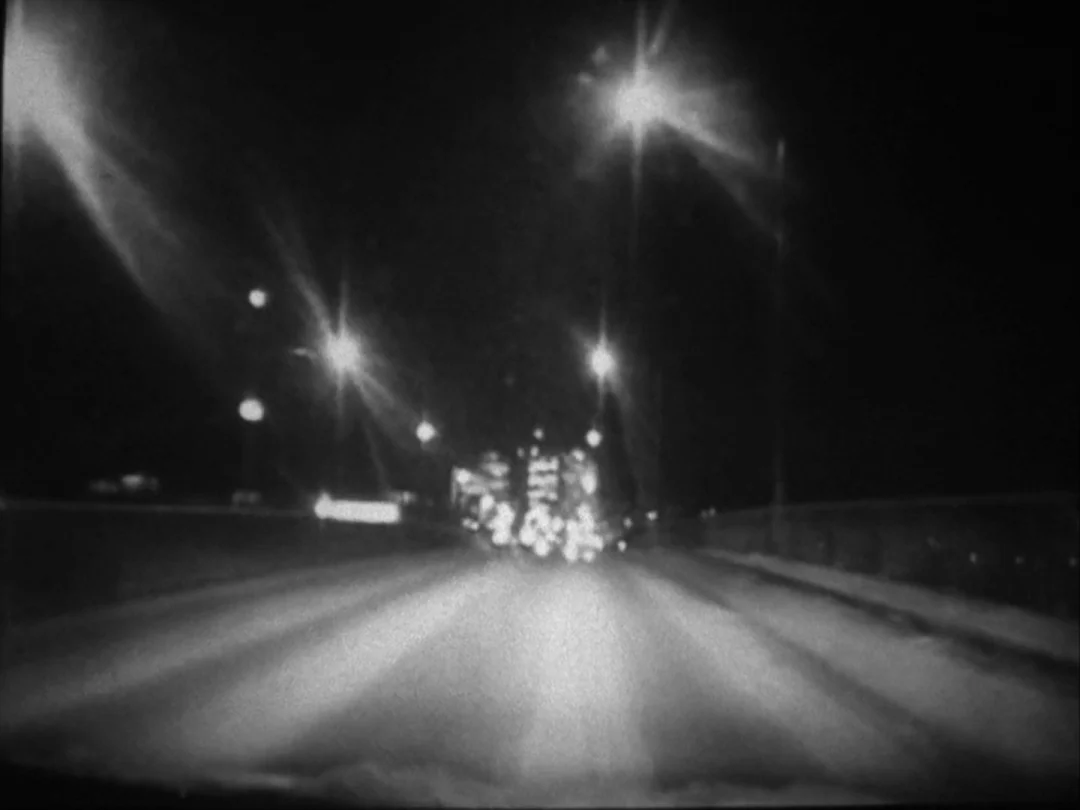 My Winnipeg (2007) movie still: wide — A dark night-time highway view captured from the perspective of a moving vehicle. Overhead streetlamps cast…