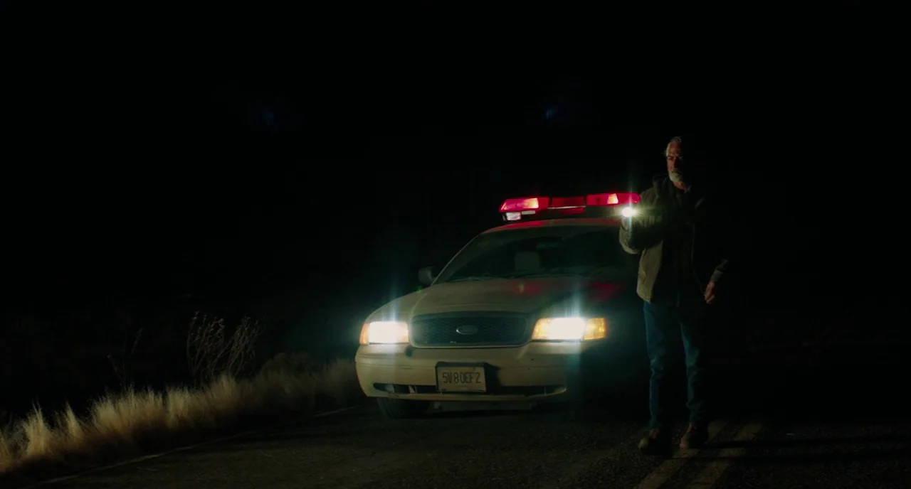 Fast Color (2018) movie still: wide — An isolated dark road at night is illuminated by a parked police vehicle. A man in work clothes walks away…