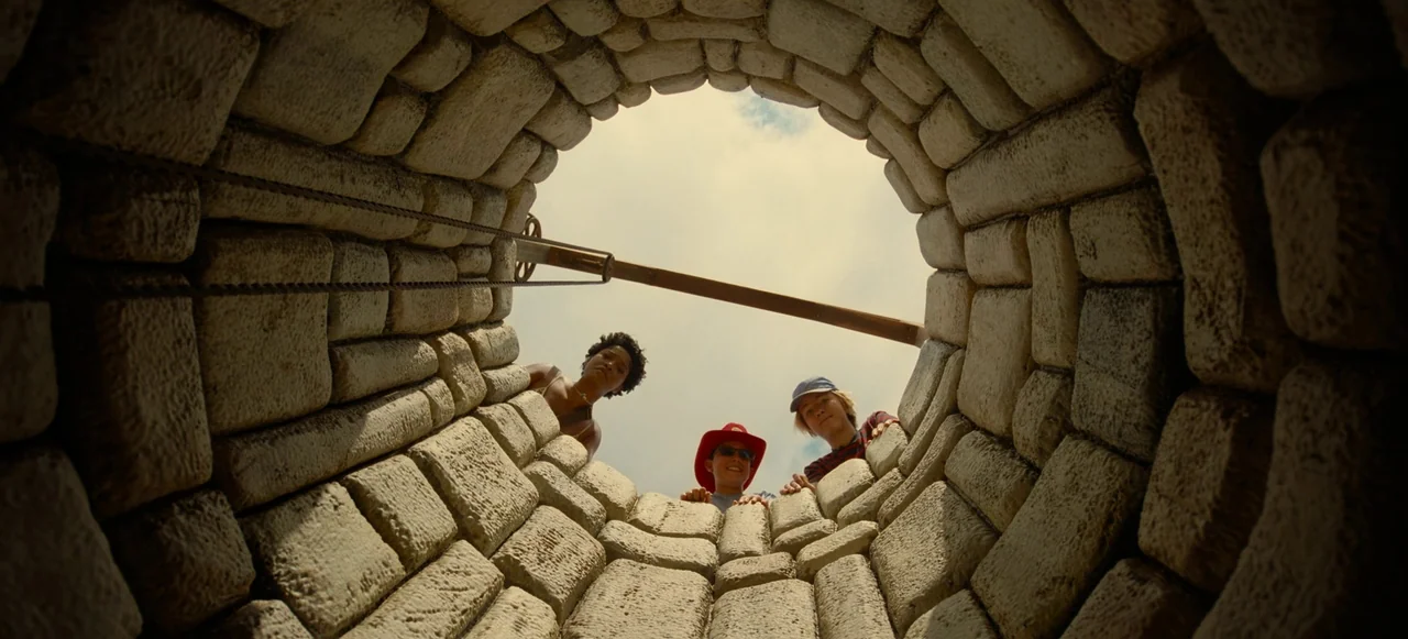 Nope (2022) movie still: wide — A stone-lined well shaft viewed from the bottom looking up toward the sky. Three teenagers peer over the circular…
