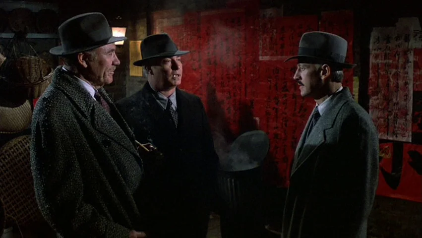 Hammett (1982) movie still: medium — Three men in fedoras converse in a dark interior space with red paper signs. A smoking metal trash can sits…