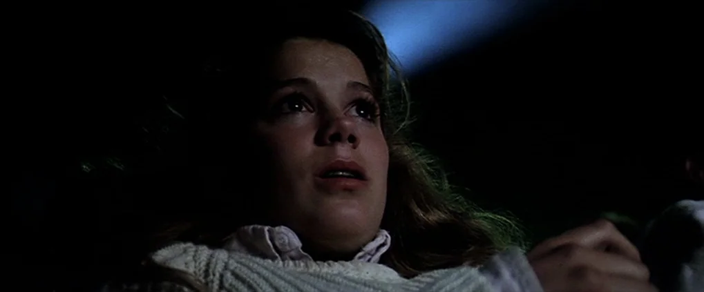 Anguish (1987) movie still: closeup — A darkened interior space shows only the upper torso and head of a young woman. The subject looks ahead with a…