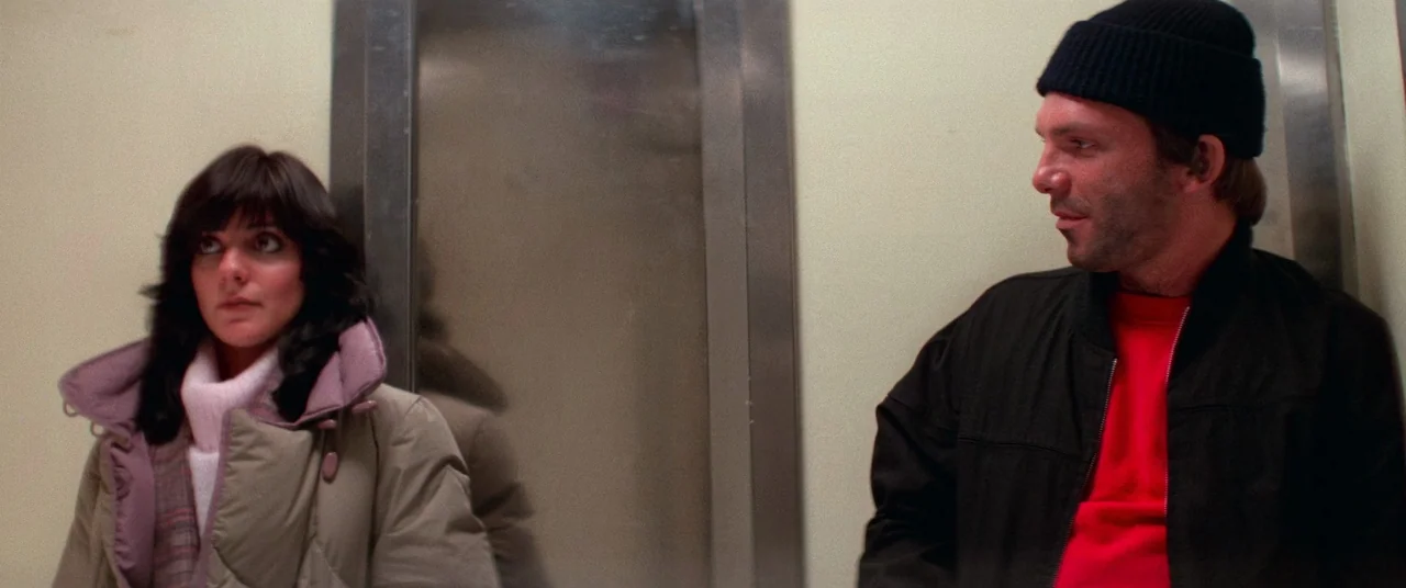 Vigilante (1982) movie still: medium — The interior of a cramped metal-lined space with beige walls and a narrow door. A man in a red sweatshirt and…