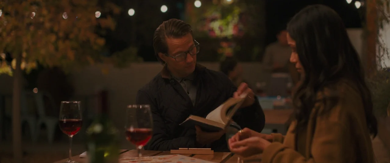 Intrusion (2021) movie still: medium — A dark outdoor patio table at night illuminated by distant string lights. A man in a quilted jacket reads a…