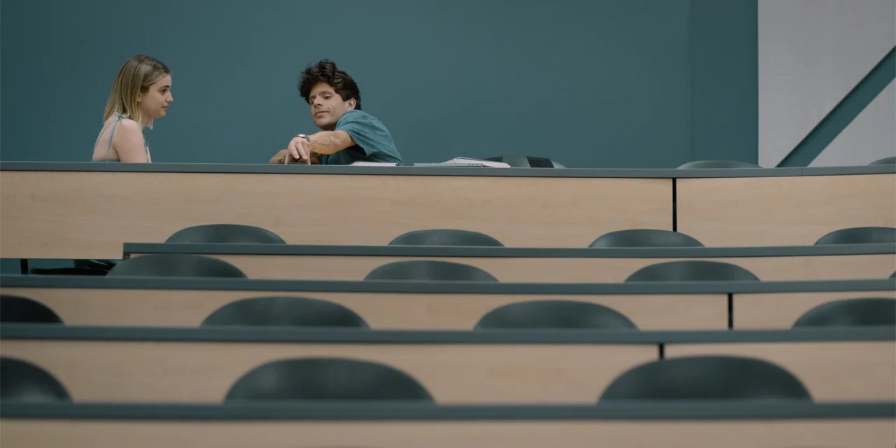 Música (2024) movie still: medium — A modern university lecture hall features rows of light wood desks and dark chairs. A young man points toward a…