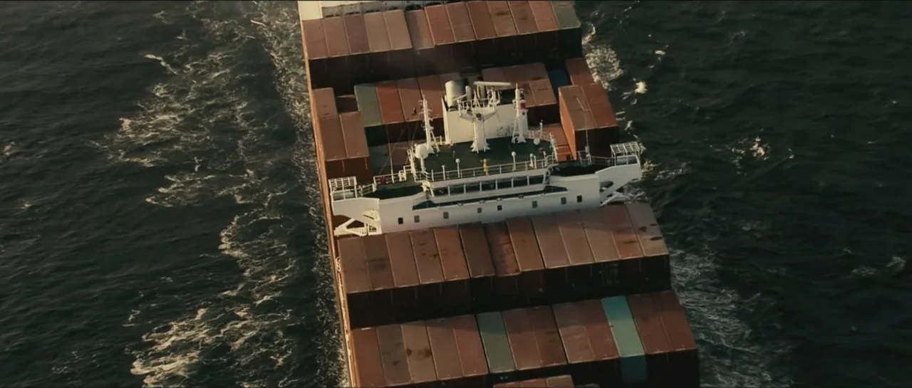 Contraband (2012) movie still: establishing — A large container ship navigates through deep water under golden hour light. The vessel is heavily…