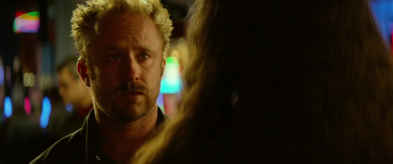 Hell or High Water (2016) movie still: closeup — A dimly lit bar setting with dark surroundings and colorful blurred neon light sources. A man with…