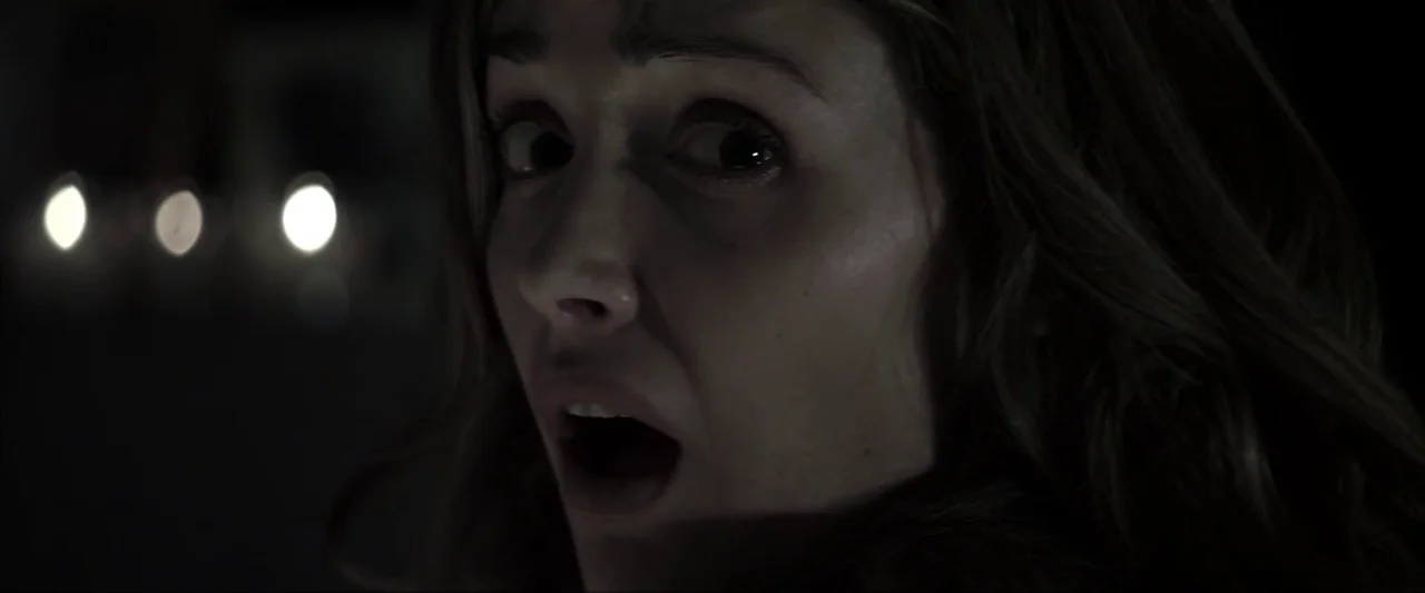 Insidious (2010) movie still: closeup — A woman is centered in a pitch-black exterior environment at night. Bright, out-of-focus headlights are…