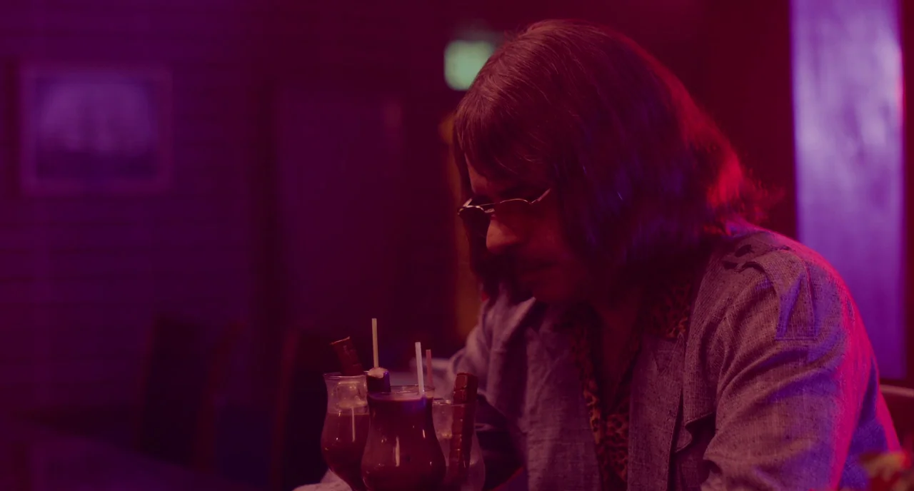 An Evening With Beverly Luff Linn (2018) movie still: medium — A dimly lit bar setting with dark wood surfaces under heavy magenta light. A man with…