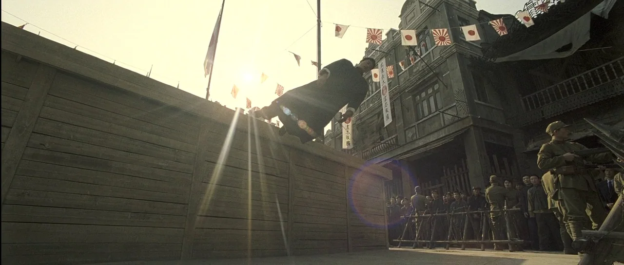 Ip Man (2008) movie still: wide — A city street decorated with flags features a large wooden barricade wall. A man jumps over the high wooden wall…