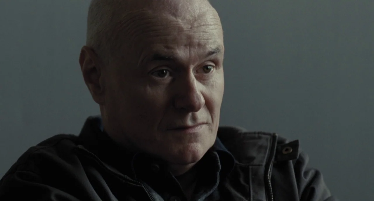 I, Daniel Blake (2016) movie still: closeup — A man stands before a uniform, neutral grey wall in a sparsely furnished space. The individual looks…