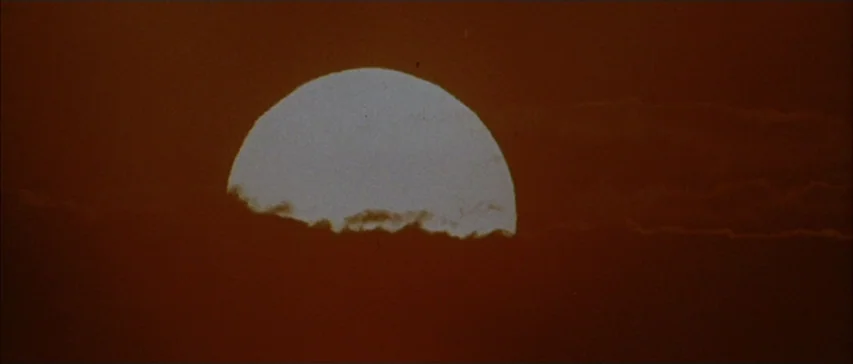 Messiah of Evil (1973) movie still: establishing — A setting sun glows bright white against a vast field of dark orange clouds. The sun is partially…