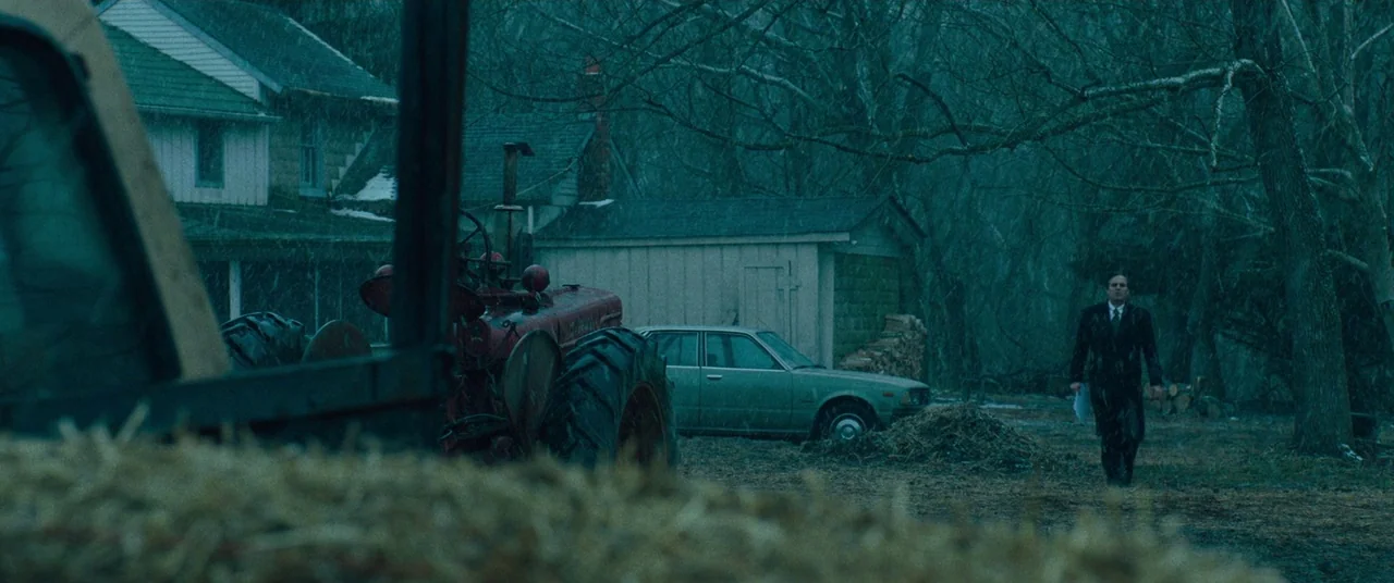 Dark Waters (2019) movie still: wide — A rural farmyard features a red tractor, an old sedan, and a residential house. A man in a suit walks across…