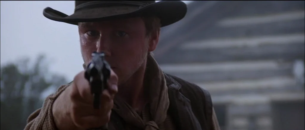 Unforgiven (1992) movie still: closeup — A man stands outdoors in a classic western setting under an overcast sky. He points a small caliber…
