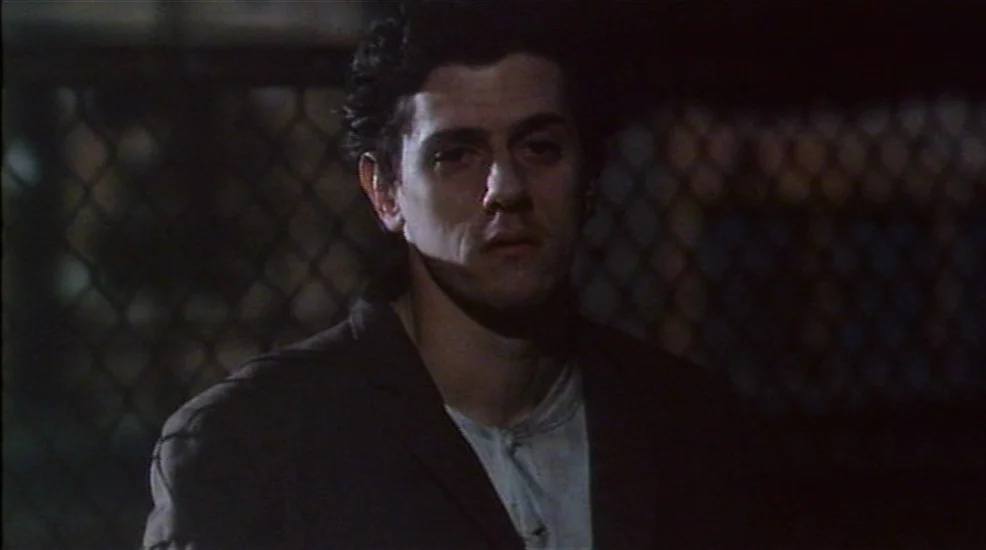 Union City (1980) movie still: medium — A man stands outdoors at night in front of a dark chain link fence. The background is out of focus and dimly…