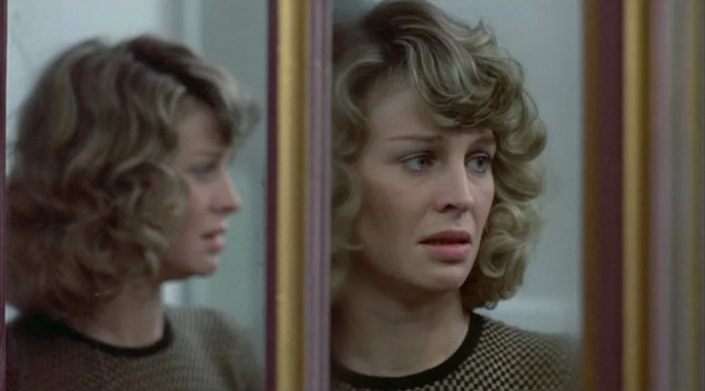 Don’t Look Now (1973) movie still: closeup — A woman looks into a mirror within a small, neutral-toned interior space. The reflection shows her side…