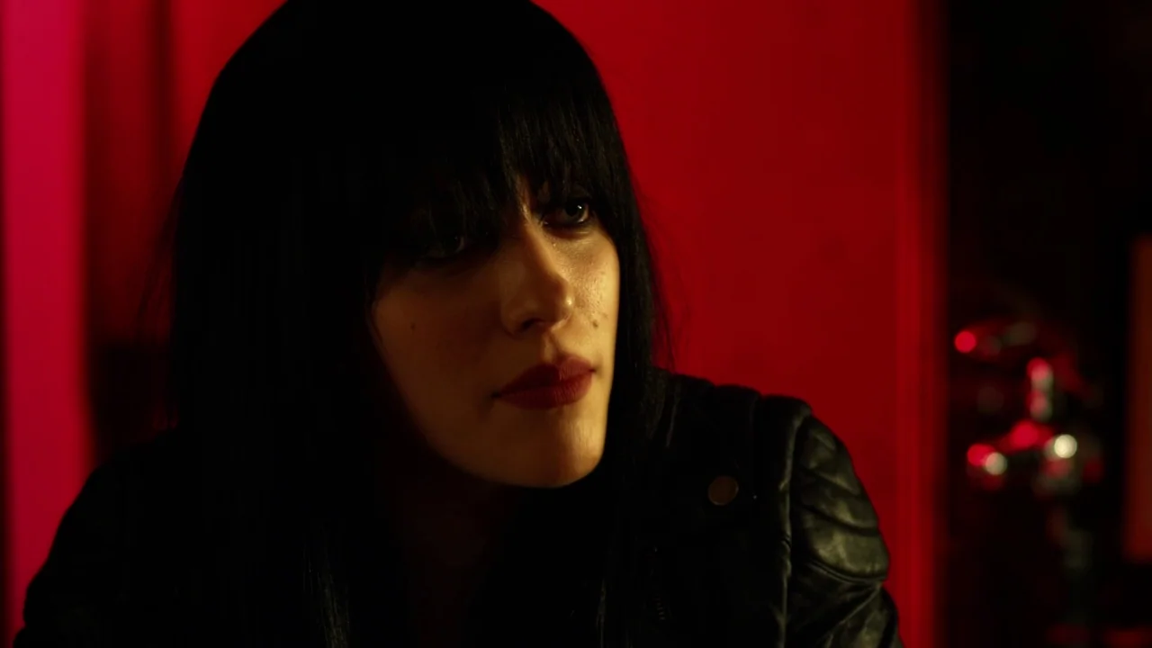 Suburban Gothic (2014) movie still: closeup — A person stands against a bright, solid red wall in a darkened room. The subject, a woman with dark…