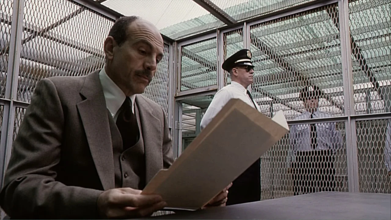 Against the Wall (1994) movie still: medium — A prison visitation room features a metal table and wire mesh dividers in the background. A man in a…