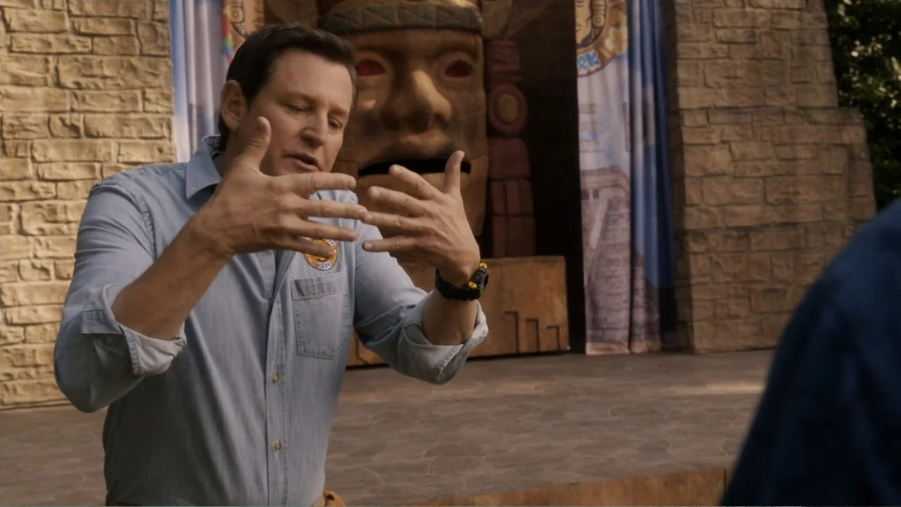 Legends of the Hidden Temple (2016) movie still: medium — A man in a light blue shirt stands in front of a stone-textured Mayan-themed facade. The…