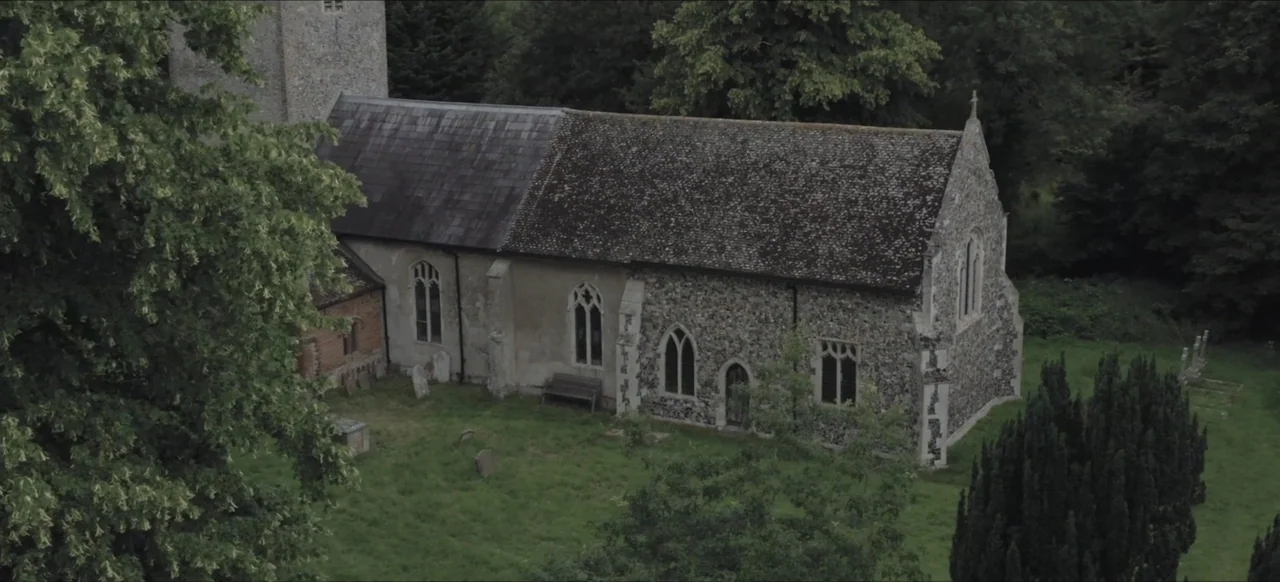 We Go In at Dawn (2020) movie still: establishing — An old stone church with a dark slate roof sits within a grassy graveyard. Several weathered…