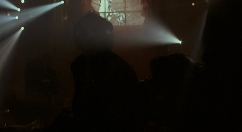 Near Dark (1987) movie still: medium — A dark, smoky space is illuminated by several harsh spotlights from an unseen source. Two silhouetted figures…