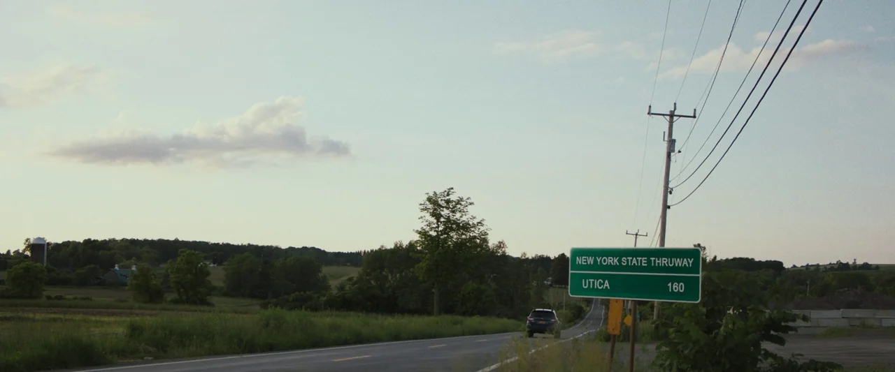 The Night House (2020) movie still: wide — A rural highway stretches through open fields and patches of trees under a sky. A green road sign…