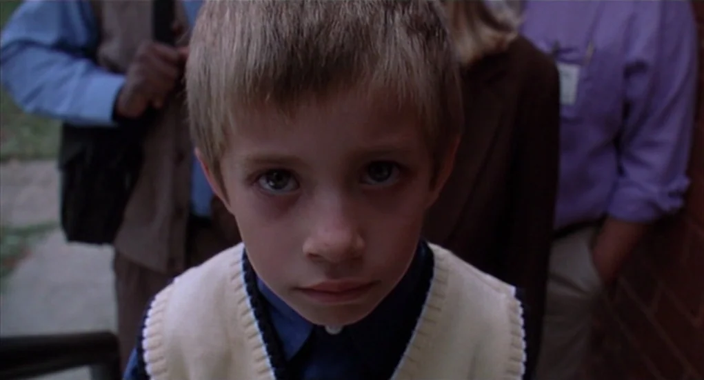 The Heart Is Deceitful Above All Things (2004) movie still: closeup — A young boy stands in the foreground with two adults blurred in the…