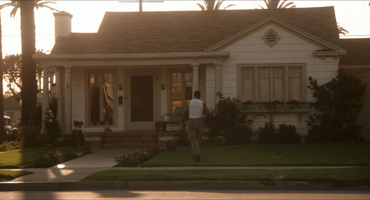 Devil In A Blue Dress (1995) movie still: wide — A single-story suburban house features white wood siding and a covered front porch. A man walks…