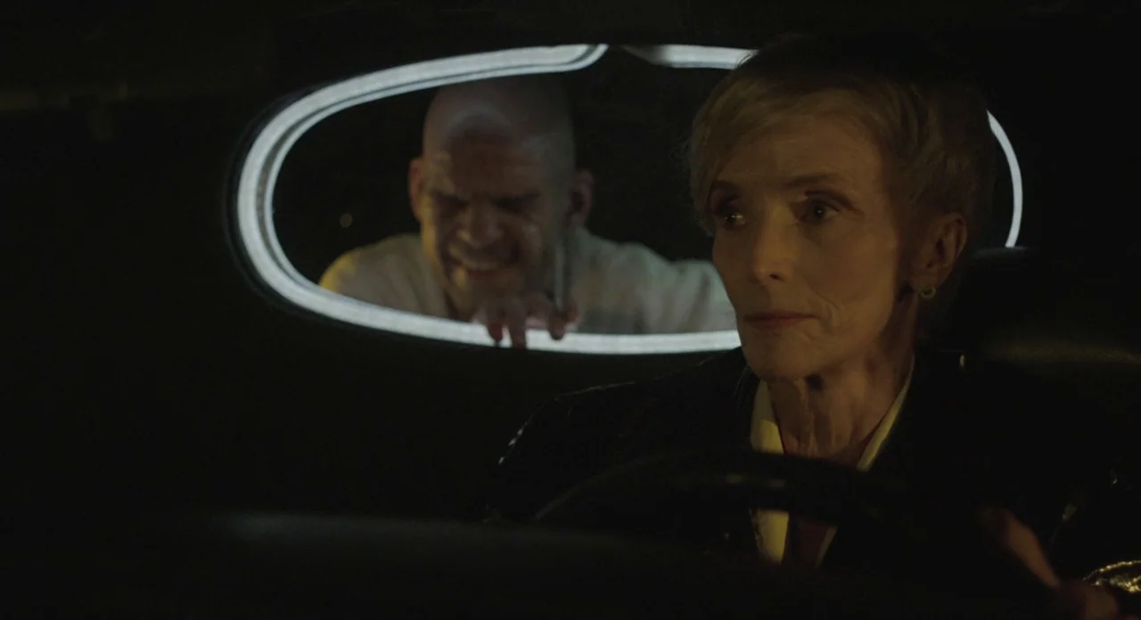 Holy Motors (2012) movie still: medium — An interior of a vehicle with dark surfaces and a circular glowing rear window. A woman sits at the…