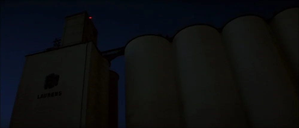 The Straight Story (1999) movie still: wide — A large industrial grain elevator stands against a dark, featureless night sky. The concrete facade…