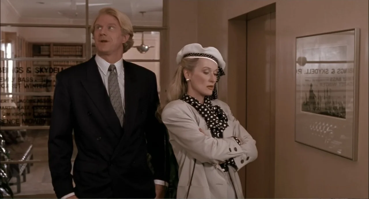 She-Devil (1989) movie still: medium — A beige office hallway with a glass-paned door and a framed print on the wall. A man and a woman stand…
