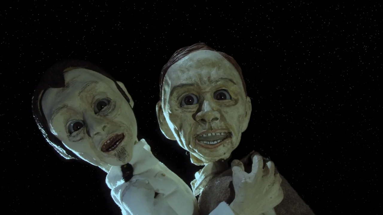 The Happiness of the Katakuris (2001) movie still: medium — Two pale, textured puppets appear floating against a dark, star-filled space. The…