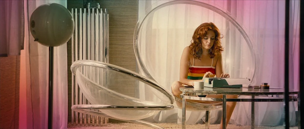 The Look of Love (2013) movie still: medium — A stylish room contains a translucent sphere chair and a low marble coffee table. A young woman with…
