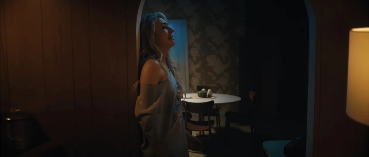 Night of the Missing (2023) movie still: medium — A dimly lit room with vintage floral wallpaper and a circular dining table set. A woman in a grey…
