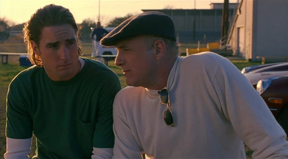Bottle Rocket (1996) movie still: medium — An outdoor field during sunset with a school building in the background. An older man wearing a cap and a…