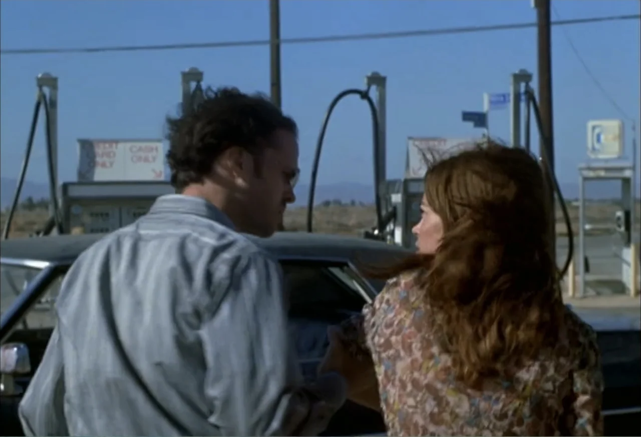 Black Cat Run (1998) movie still: medium — An outdoor gas station lot under a clear blue sky with fuel pumps visible. A man and a woman stand by a…