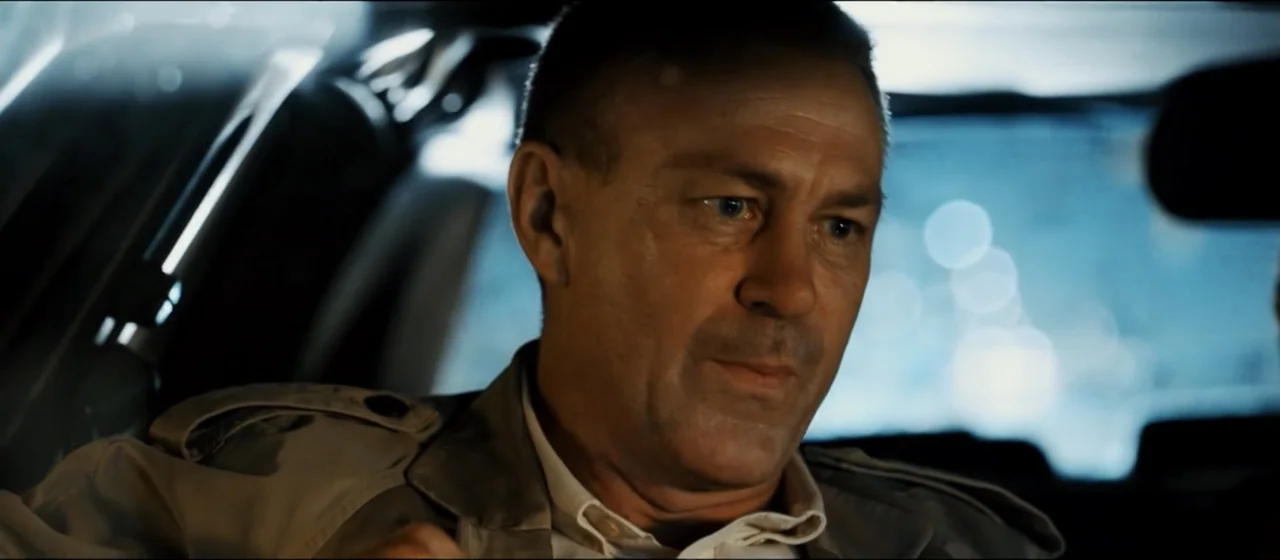 Guns Akimbo (2020) movie still: closeup — A man sits in the passenger seat of a car at night with city lights blurred. He gazes forward while the…