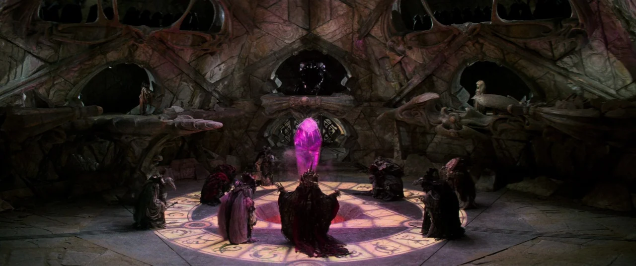 The Dark Crystal (1982) movie still: wide — A dark subterranean stone chamber features circular alcoves built into the rock walls. Several shrouded,…