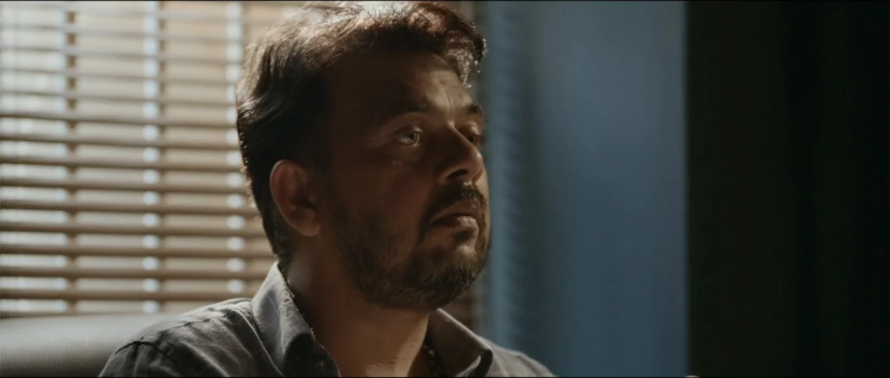 Garudan (2024) movie still: closeup — An interior office space features horizontal window blinds providing backlighting for the subject. A man sits…