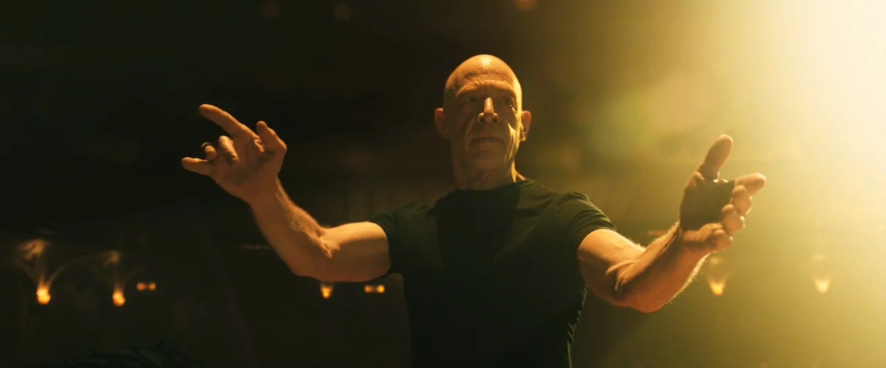 Whiplash (2014) movie still: medium — A dimly lit concert hall interior with golden stage lights behind the subject. A bald man wearing a black…